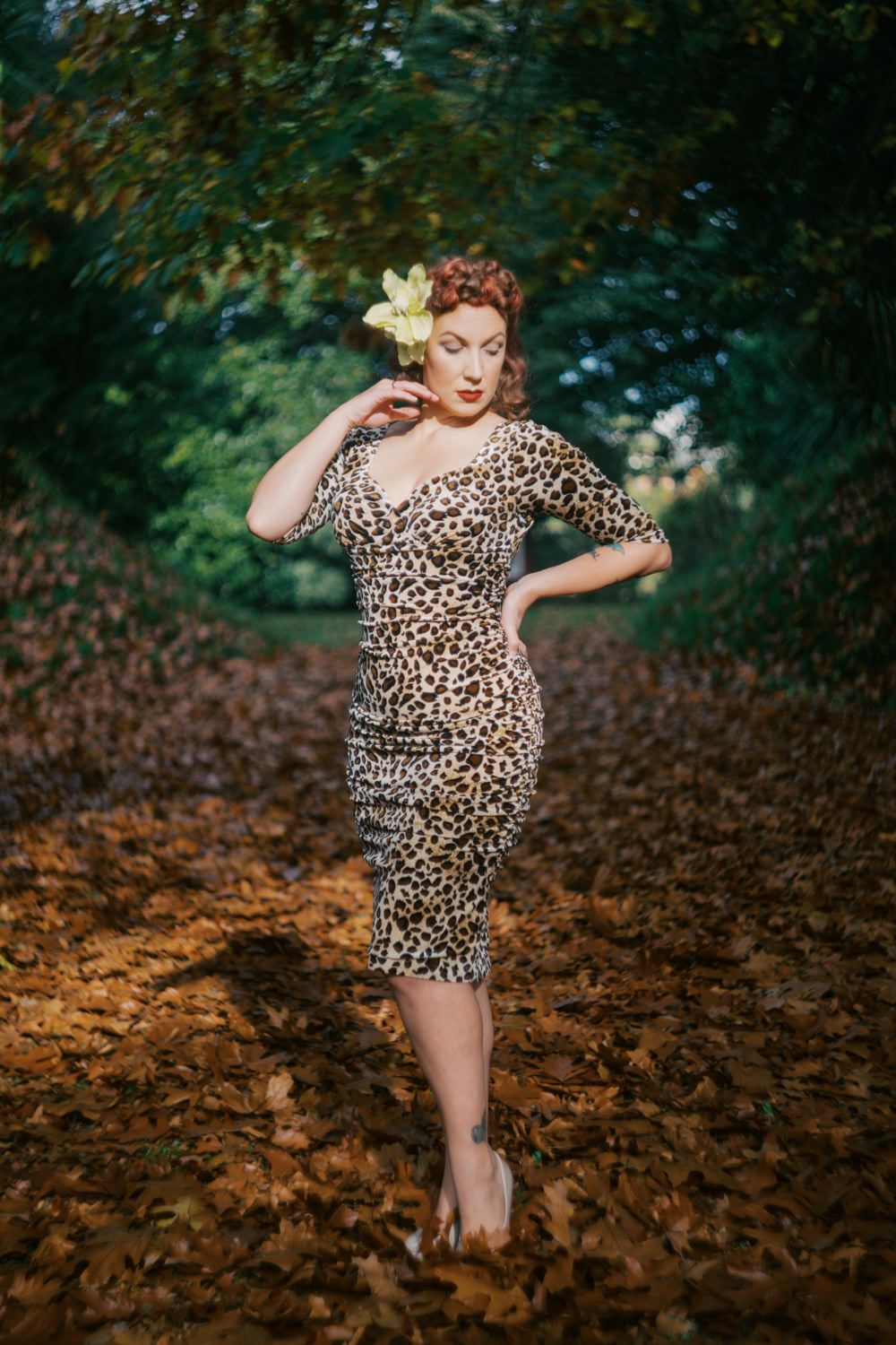 Woman in leopard print dress with yellow flower hair accessory poses among autumn leaves in a lush green tree-lined path.
