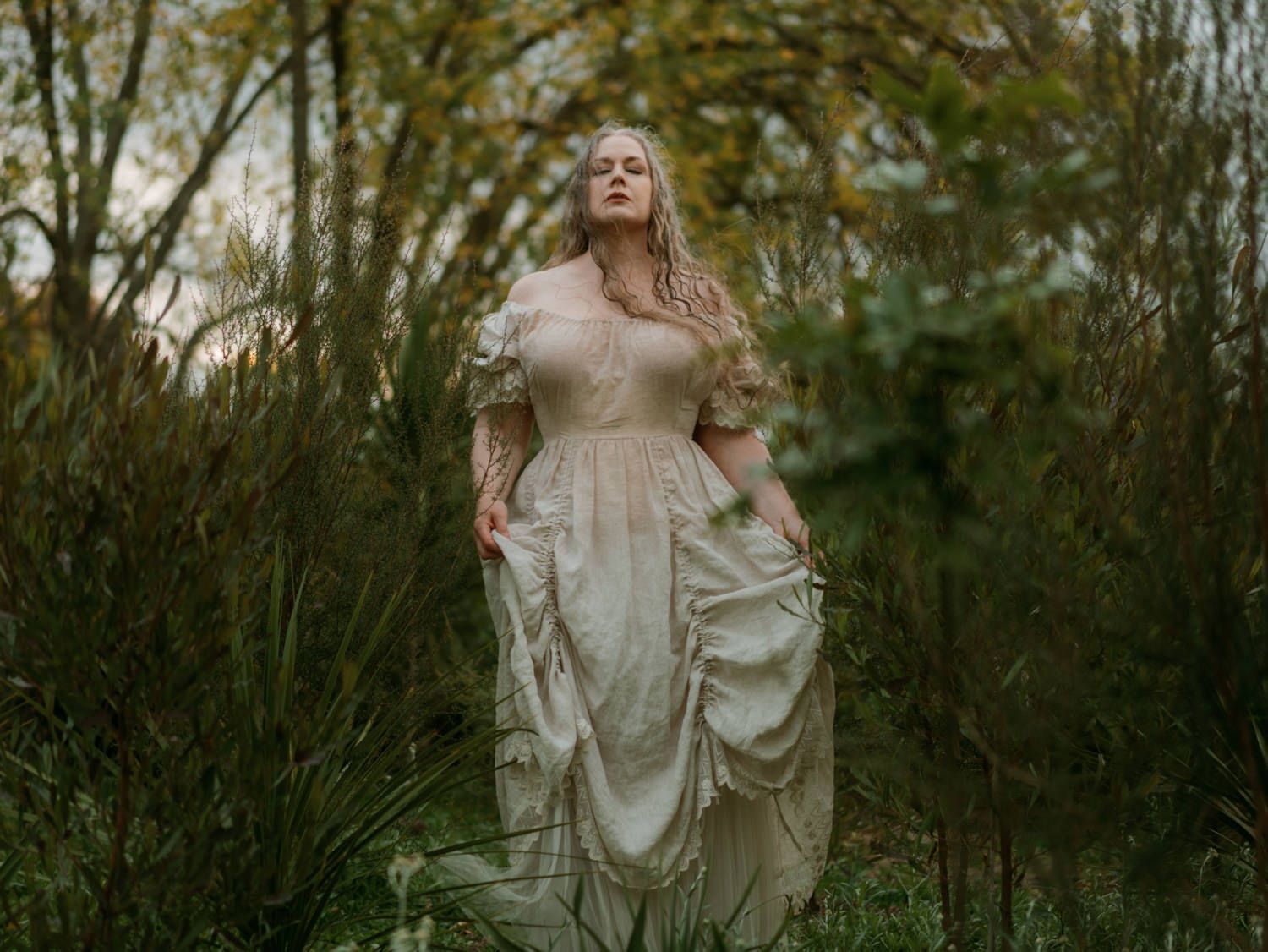 Woman in vintage cream gown standing among lush green trees in a dreamy outdoor forest setting.