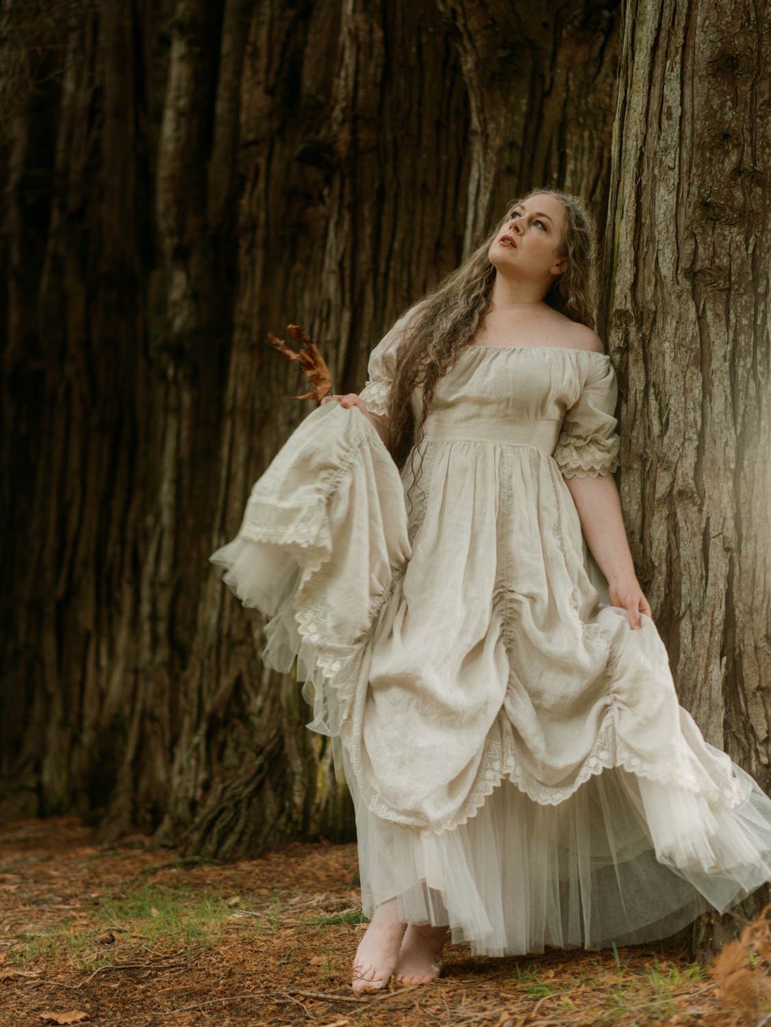 Woman in elegant off-shoulder ivory gown poses gracefully against a large redwood tree in a forest setting.
