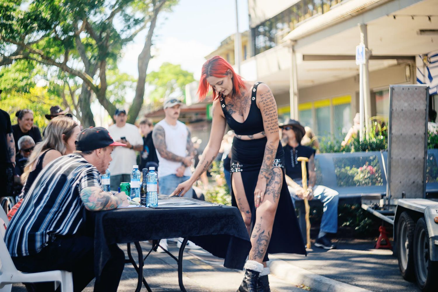 Heavily tattooed woman with red hair leans over outdoor table at a sunny street event with crowd in background.