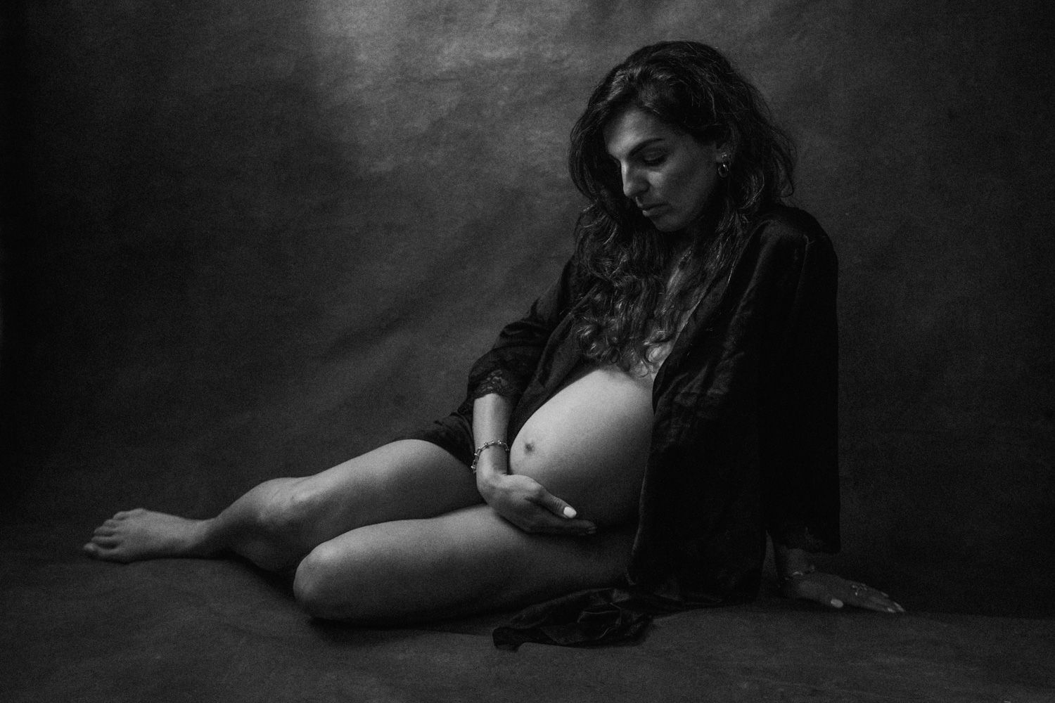 Black and white maternity photo of a pregnant woman sitting on the floor cradling her baby bump.