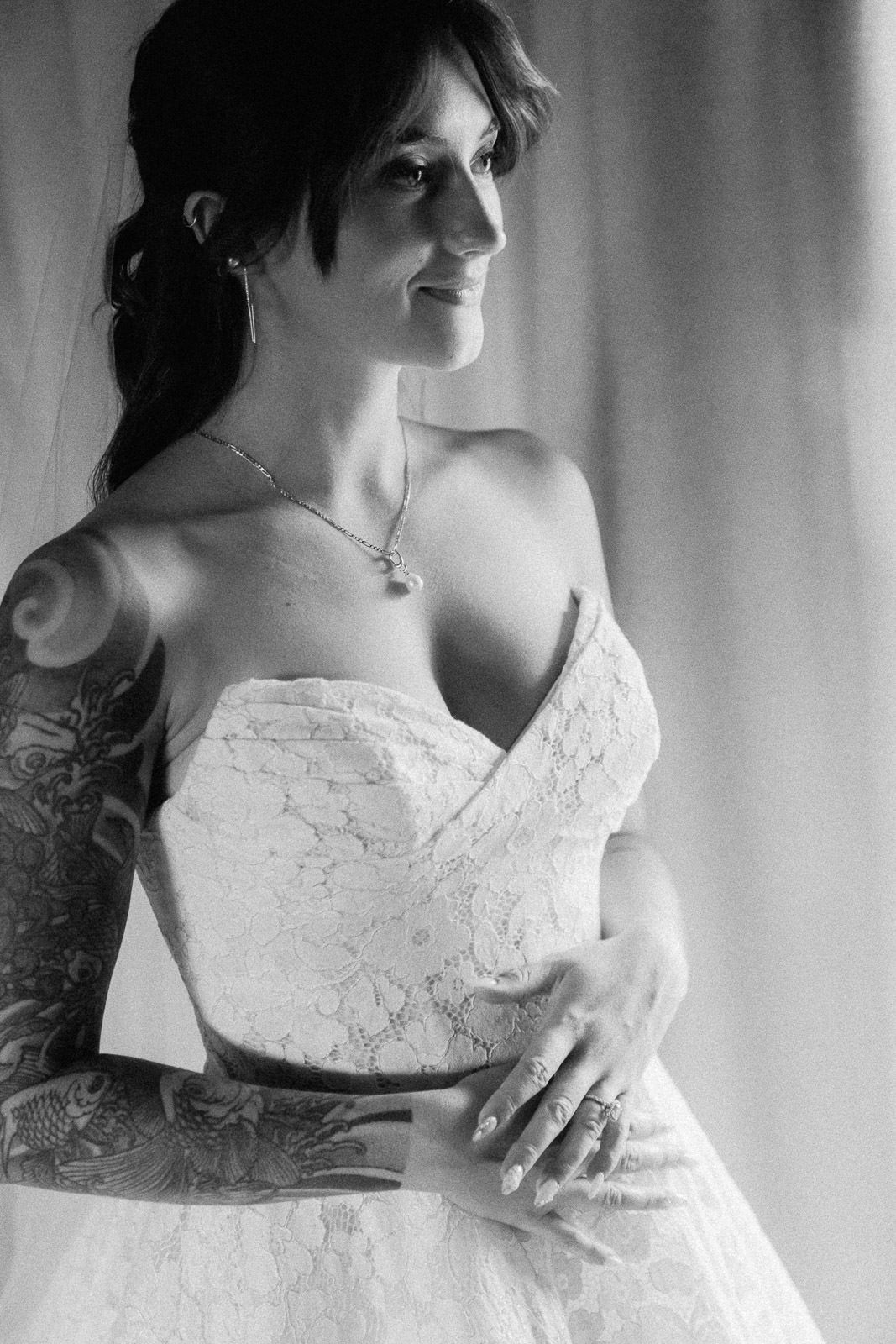 Black and white photo of a tattooed bride in a strapless lace wedding gown, smiling softly near a window.