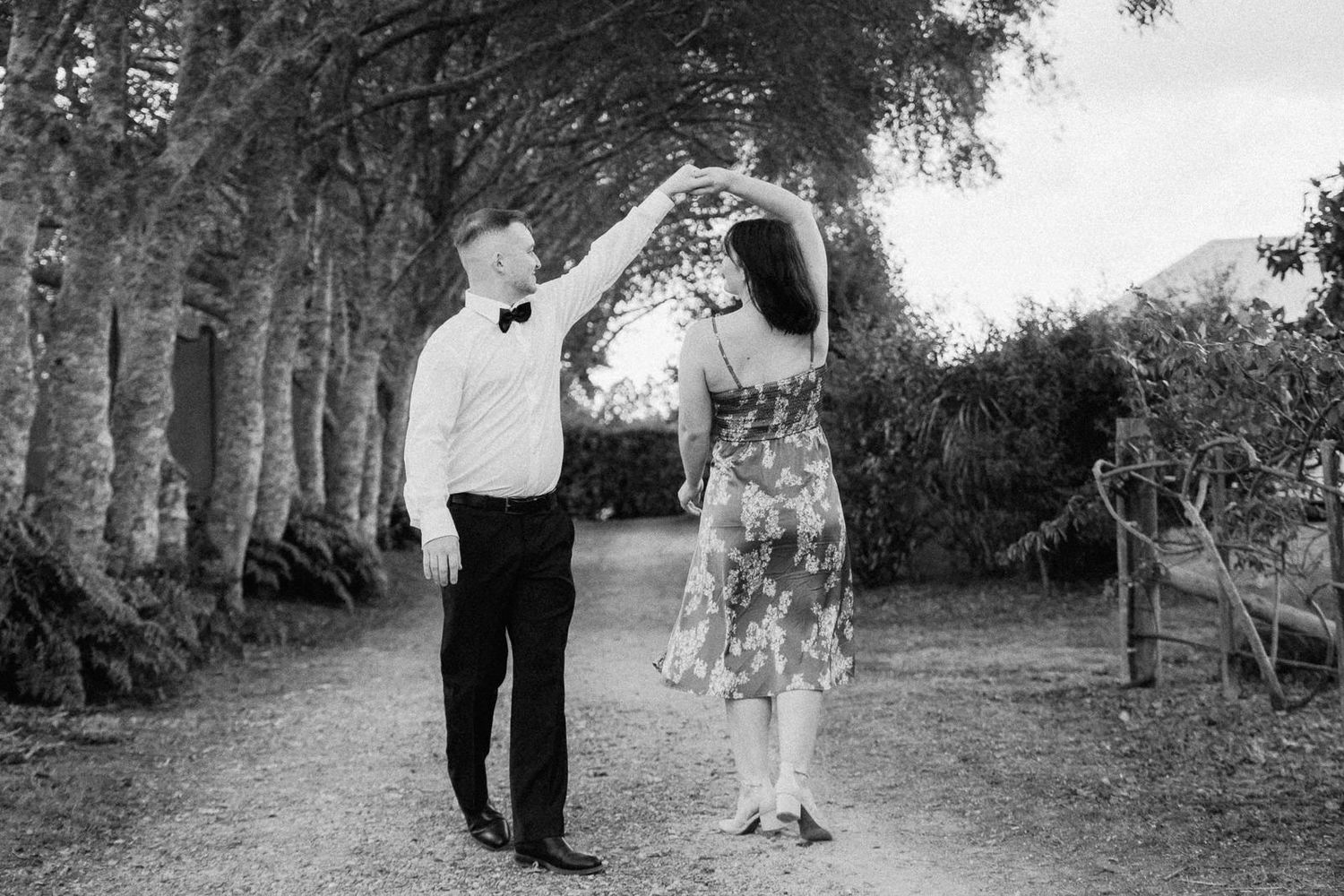 Couple dancing outdoors on a garden path lined with trees in a black and white wedding photo.