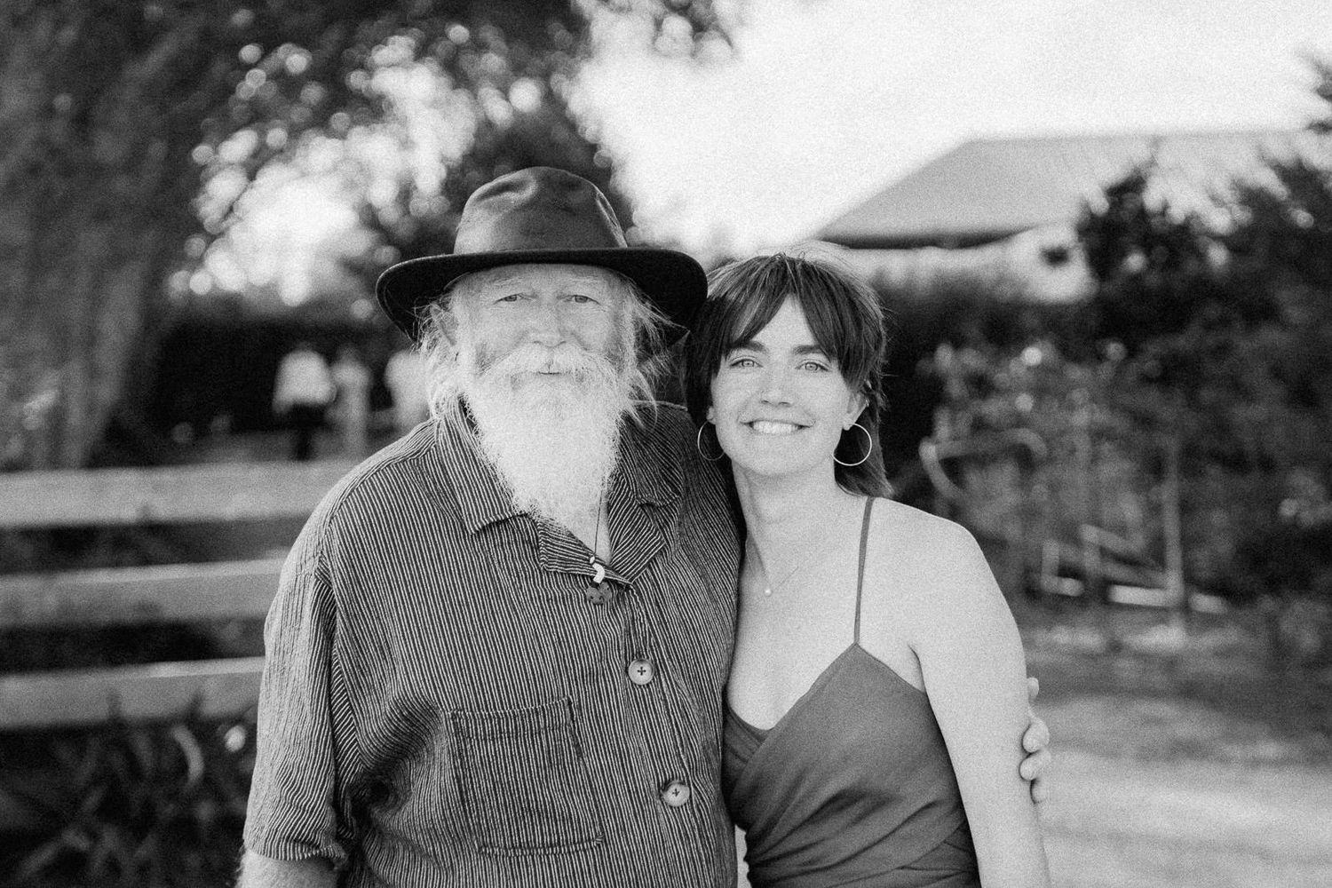 Black and white photo of an elderly bearded man in a hat and a smiling woman posing together outdoors.