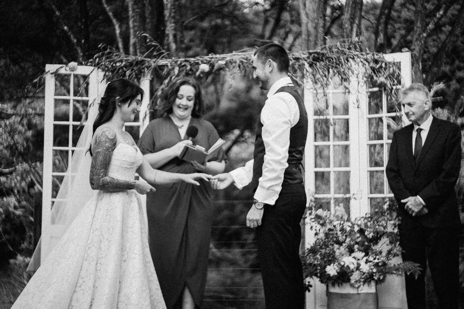 Black and white wedding ceremony photo of bride and groom exchanging rings outdoors with officiant and guest.