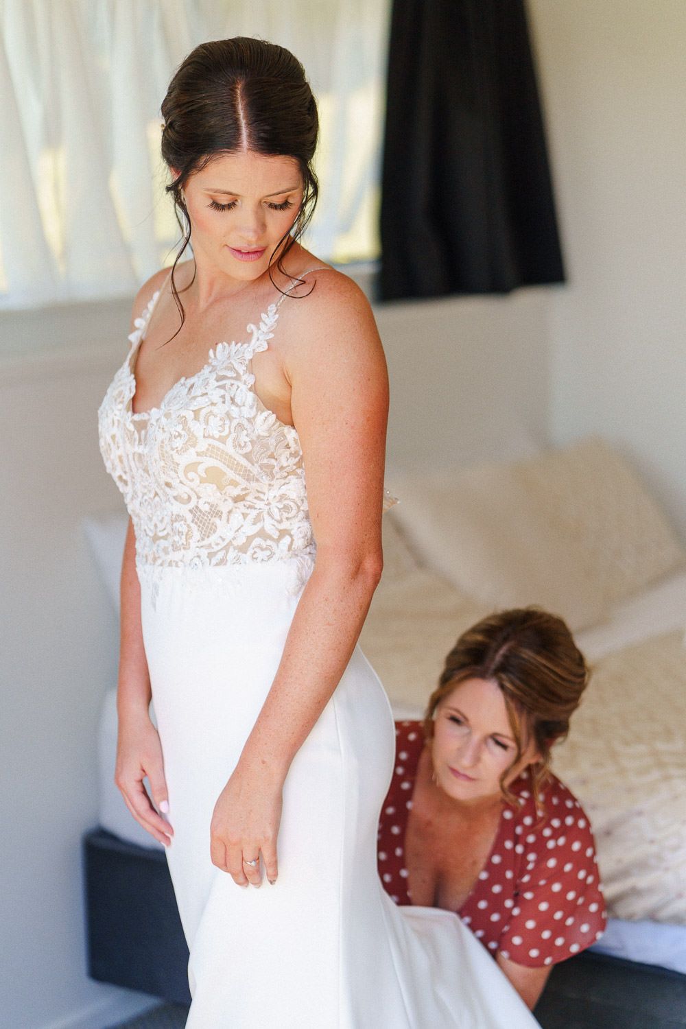 Bride in white lace wedding gown being helped by bridesmaid in red polka dot dress during bridal preparations.