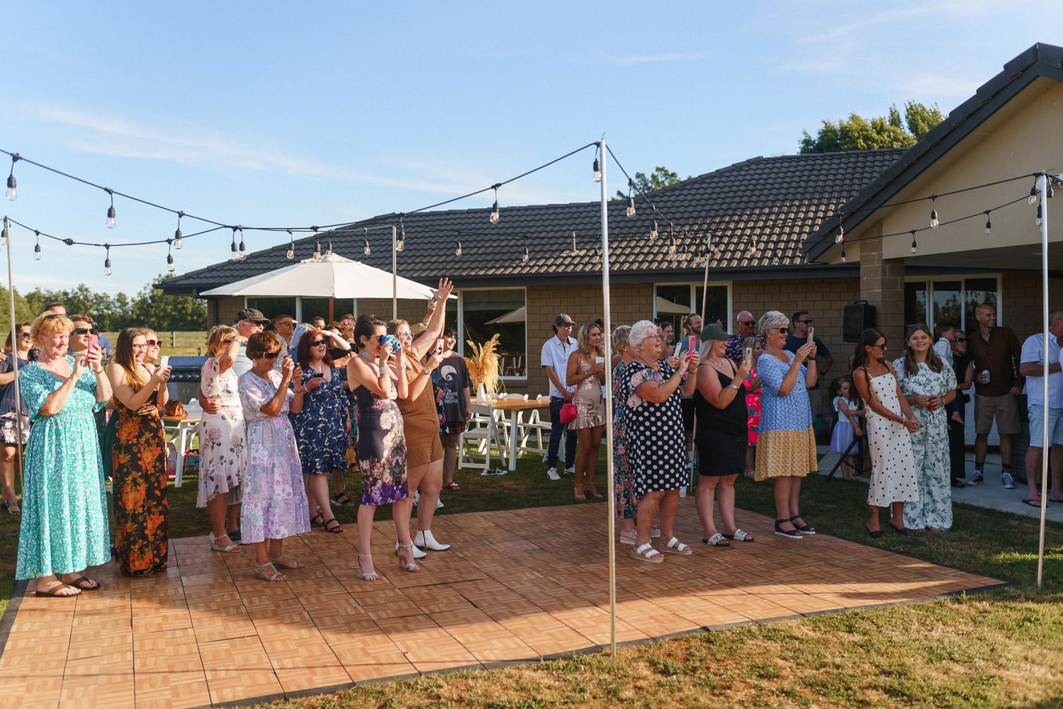 Guests clapping and celebrating outdoors at a backyard party with string lights and a modern house.