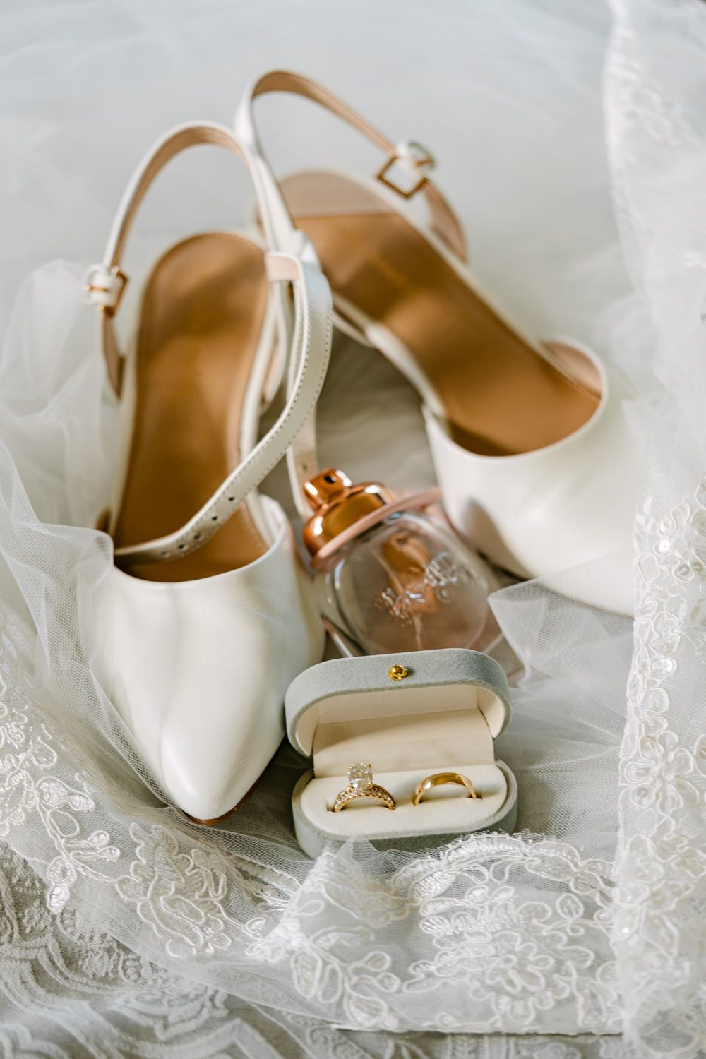 Elegant white bridal heels with ankle straps, gold wedding rings in a velvet box, and perfume on lace veil.