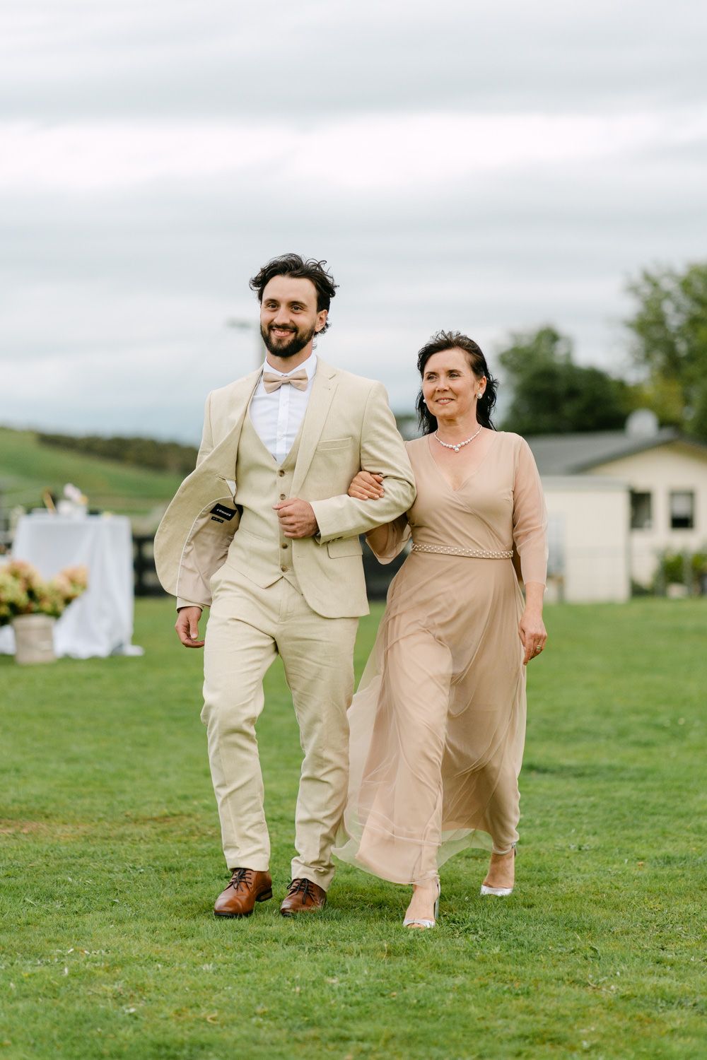 A couple in elegant beige attire walks hand-in-hand on green grass at an outdoor wedding venue.