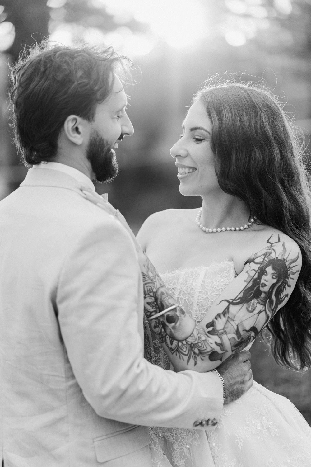 Black and white wedding photo of smiling tattooed bride and bearded groom gazing at each other outdoors.