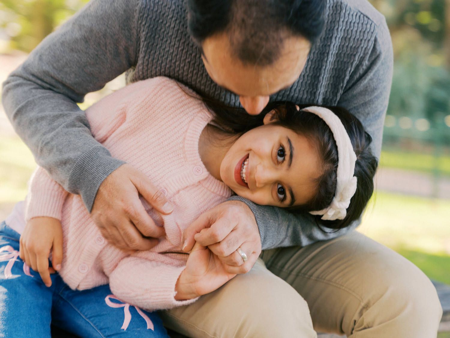 A parent lovingly hugs a smiling young girl in a pink sweater outdoors in a park setting.