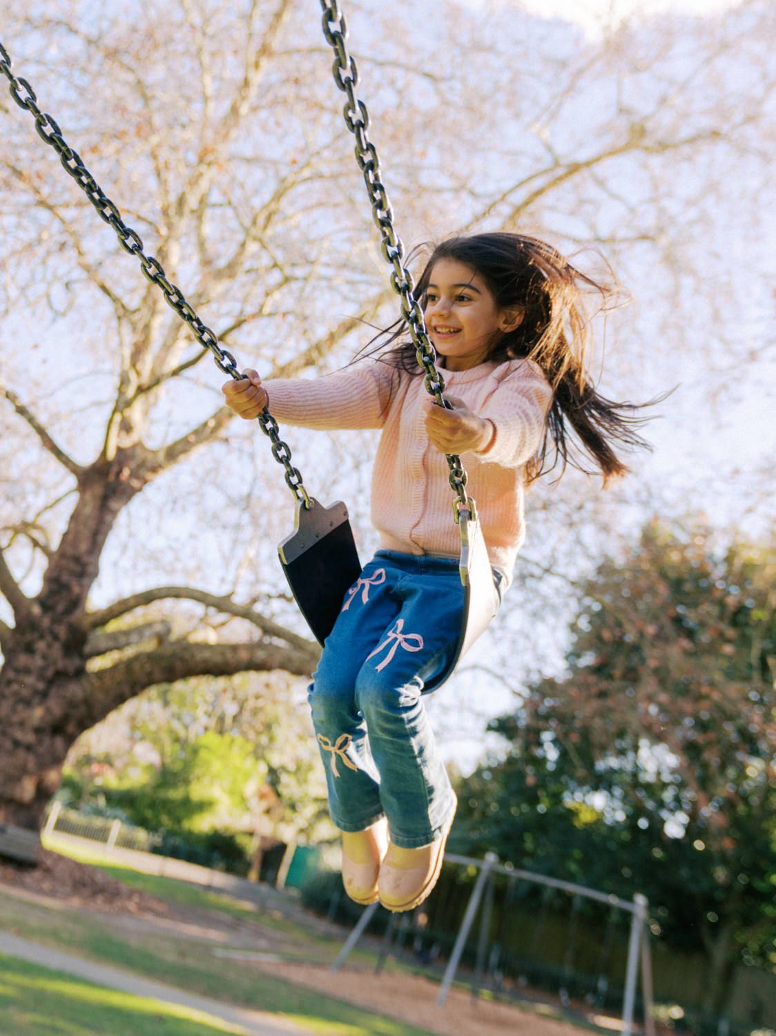 Happy girl swinging high on a chain swing in a park, wearing pink hoodie and blue jogger pants.
