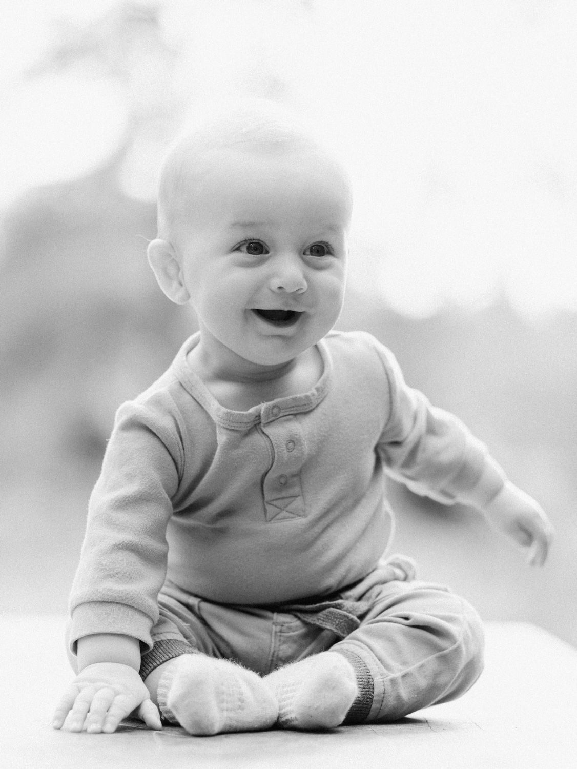 Black and white photo of a happy smiling baby sitting upright wearing a casual onesie outfit.