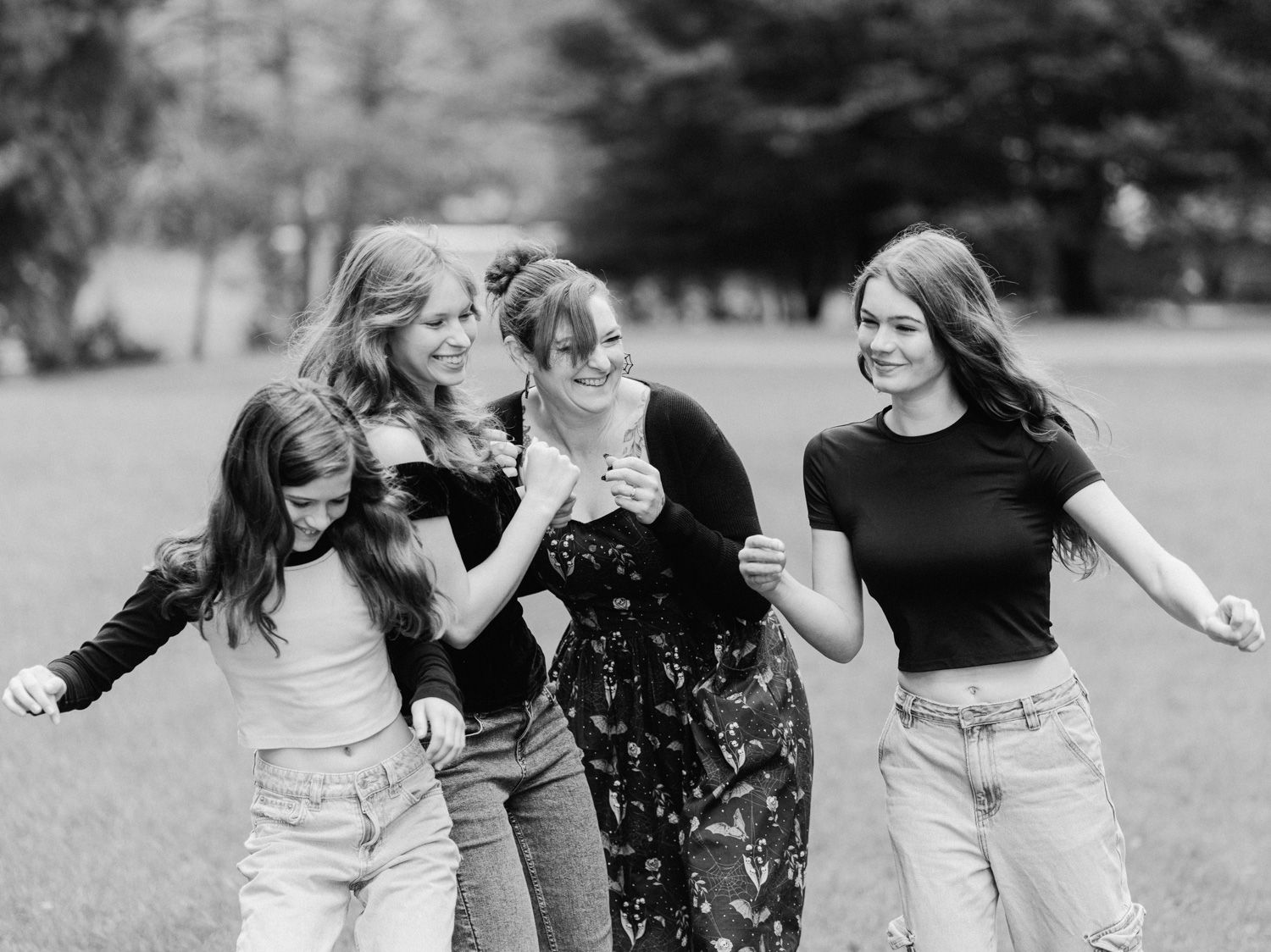 Five young women laughing and dancing together in a park, captured in a joyful black and white outdoor photo.