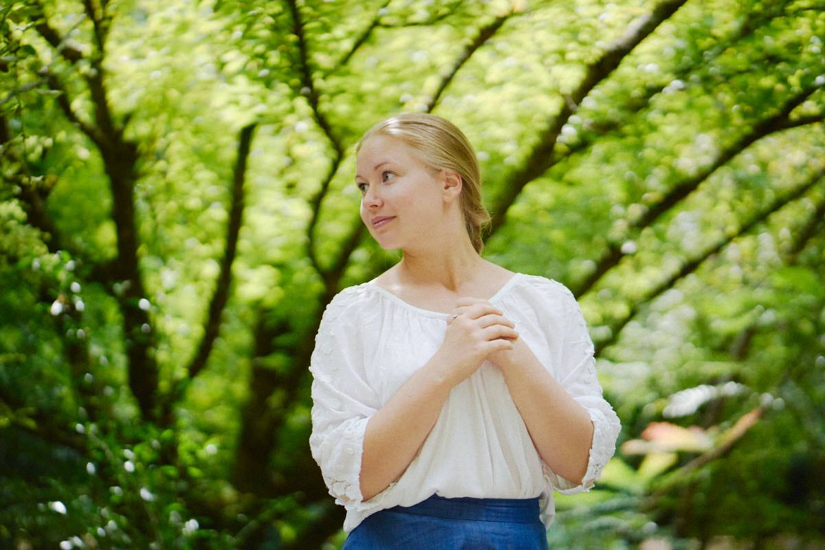 Young blonde woman in white blouse stands among lush green trees, hands clasped, gazing thoughtfully to the side.