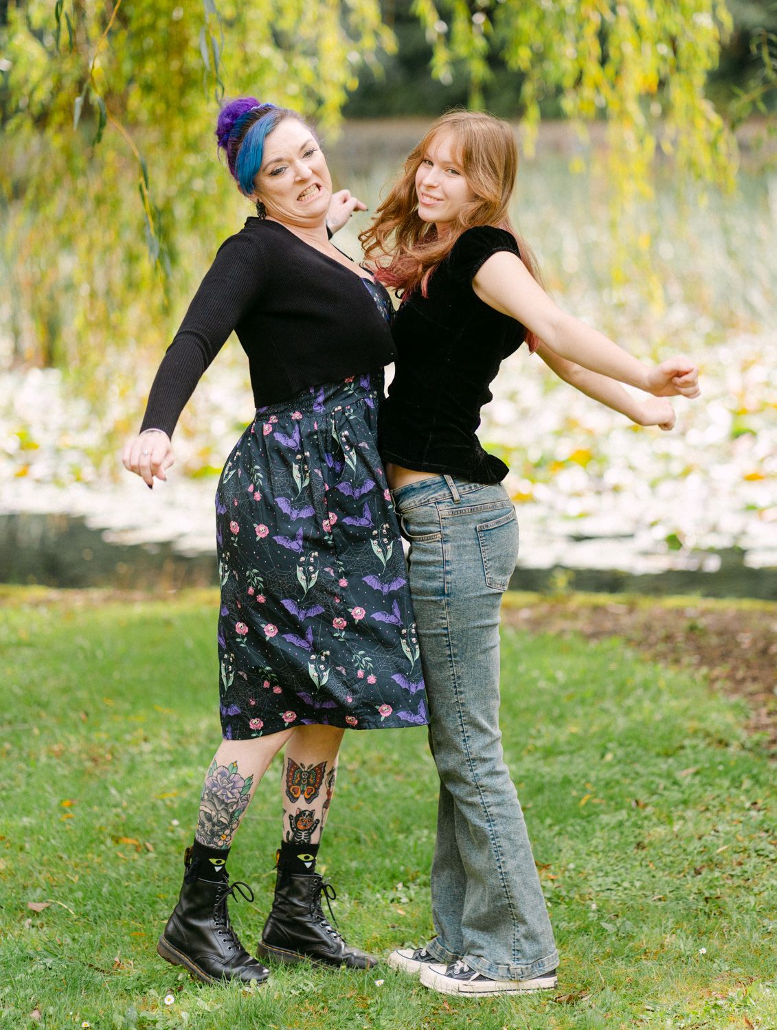 Two women dancing outdoors near a pond, one with blue hair and tattoos in a floral skirt, the other in jeans.