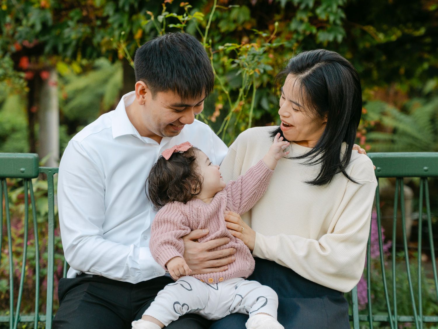 A young Asian family shares a tender moment outdoors, with parents smiling at their baby girl in a pink sweater.
