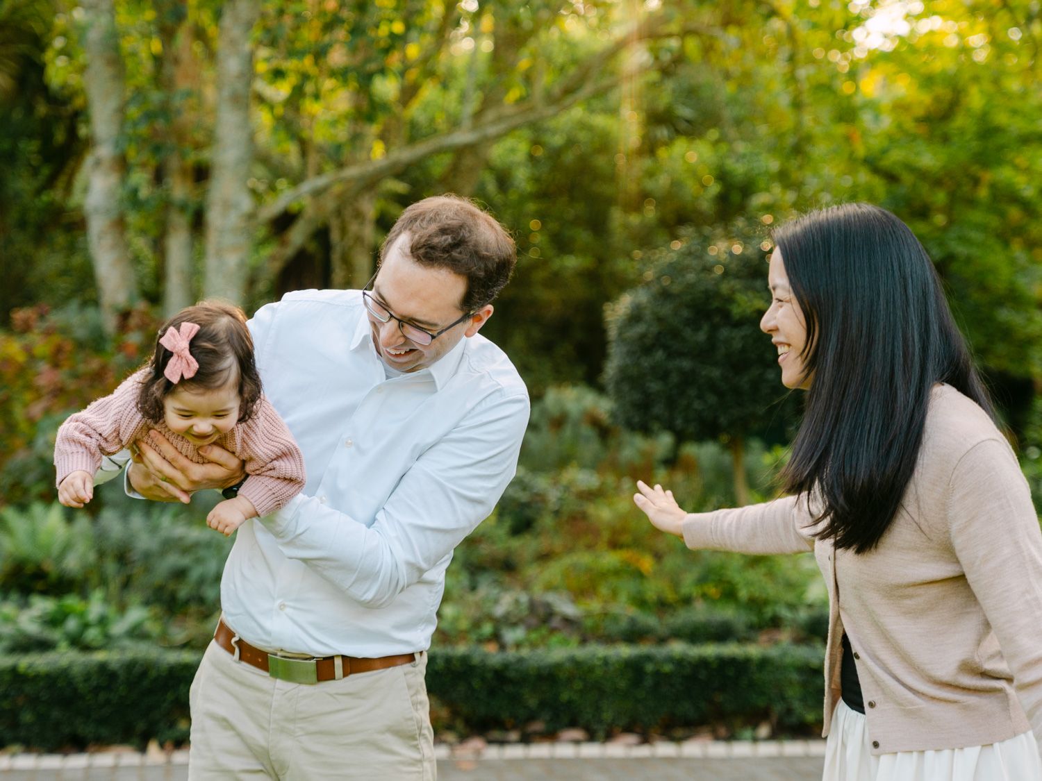 Family of three enjoying playful moment outdoors, father holding toddler girl with pink bow, mother laughing in garden.