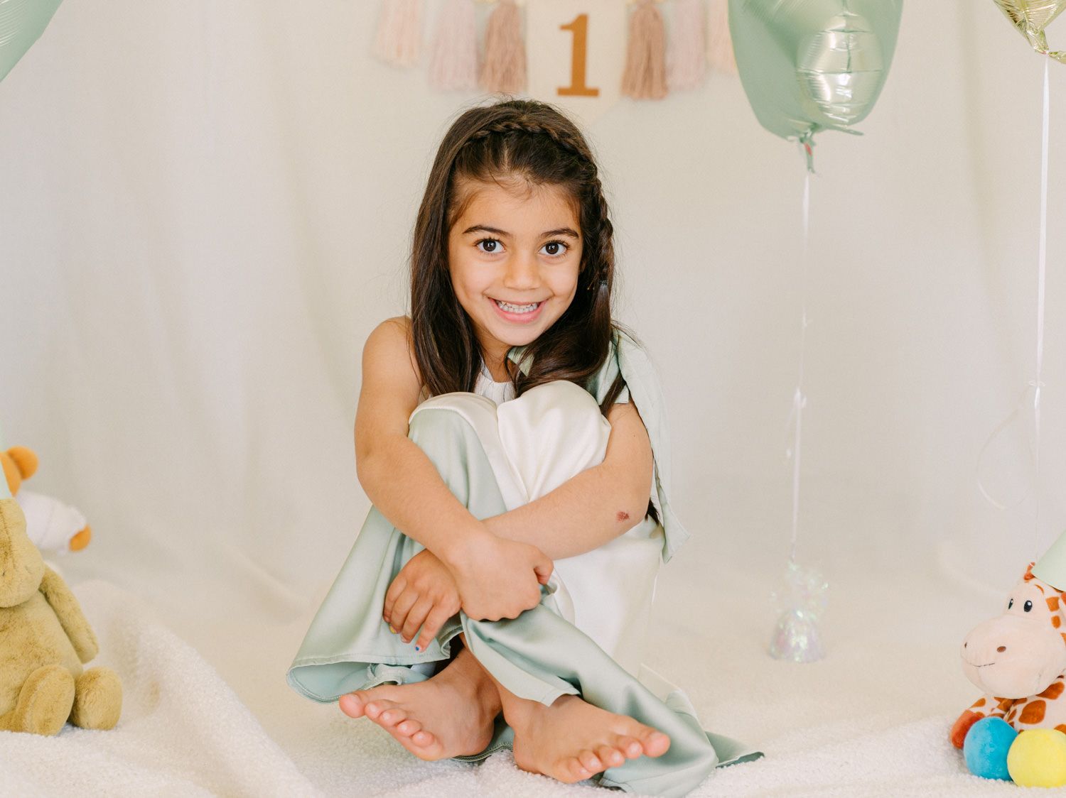 A smiling young girl sits cross-legged on white fabric surrounded by balloons and toys at a first birthday party celebration.