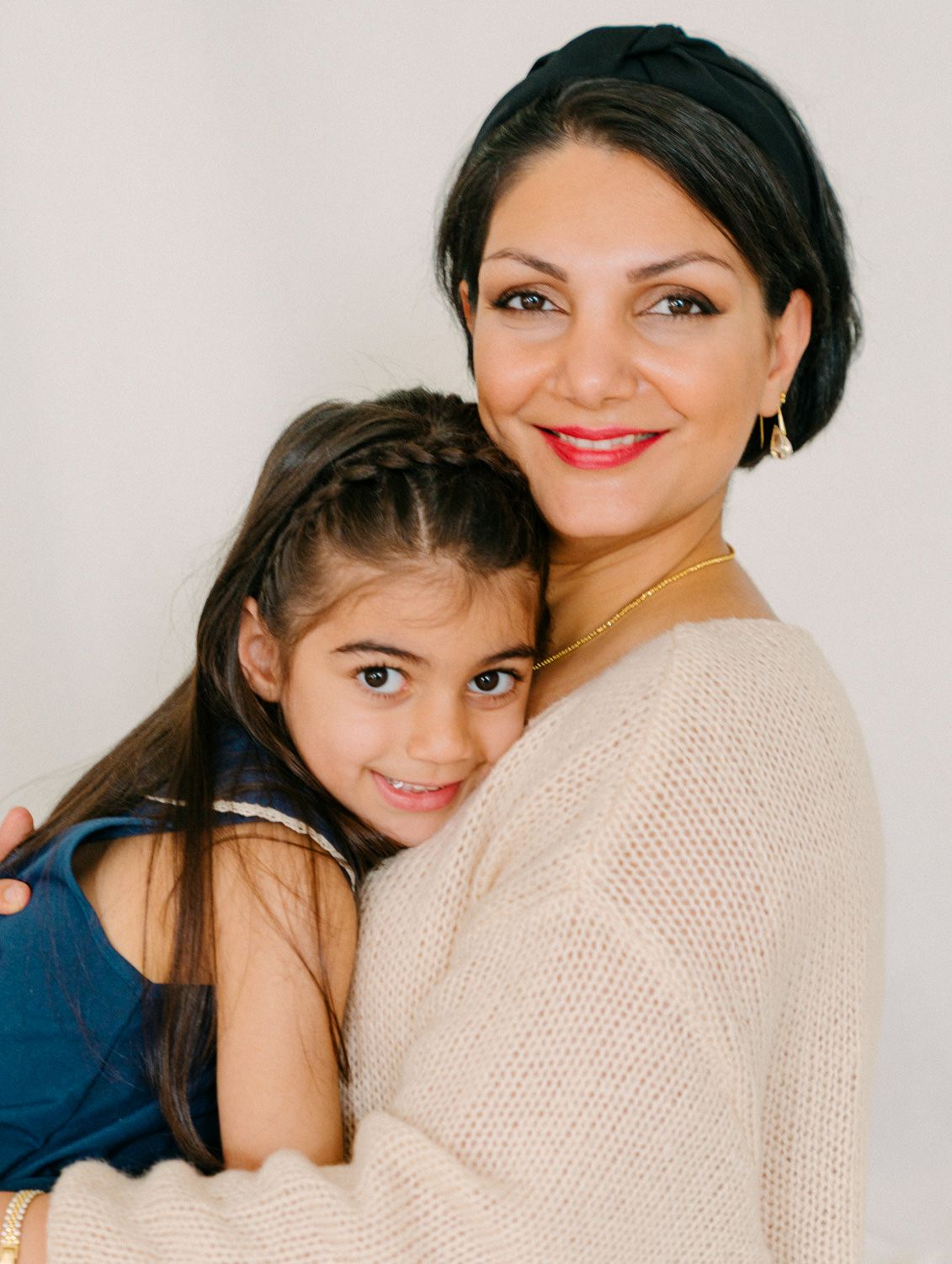 A woman in a beige sweater and headband smiles warmly while hugging a young girl with braided hair.