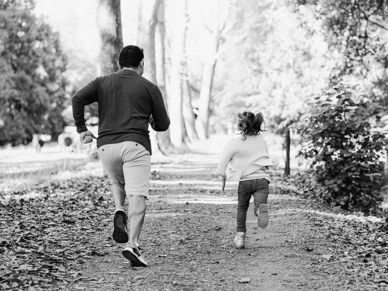 Adult and child running together on a tree-lined park path, captured in black and white from behind.