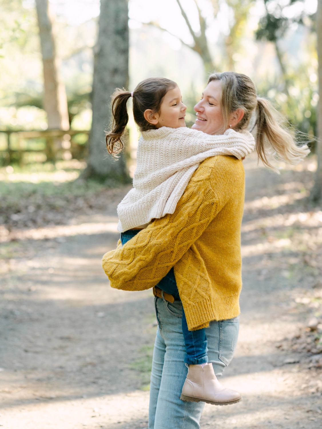 A mother in a yellow sweater joyfully lifts her smiling daughter in a cream knit sweater outdoors on a sunny path.