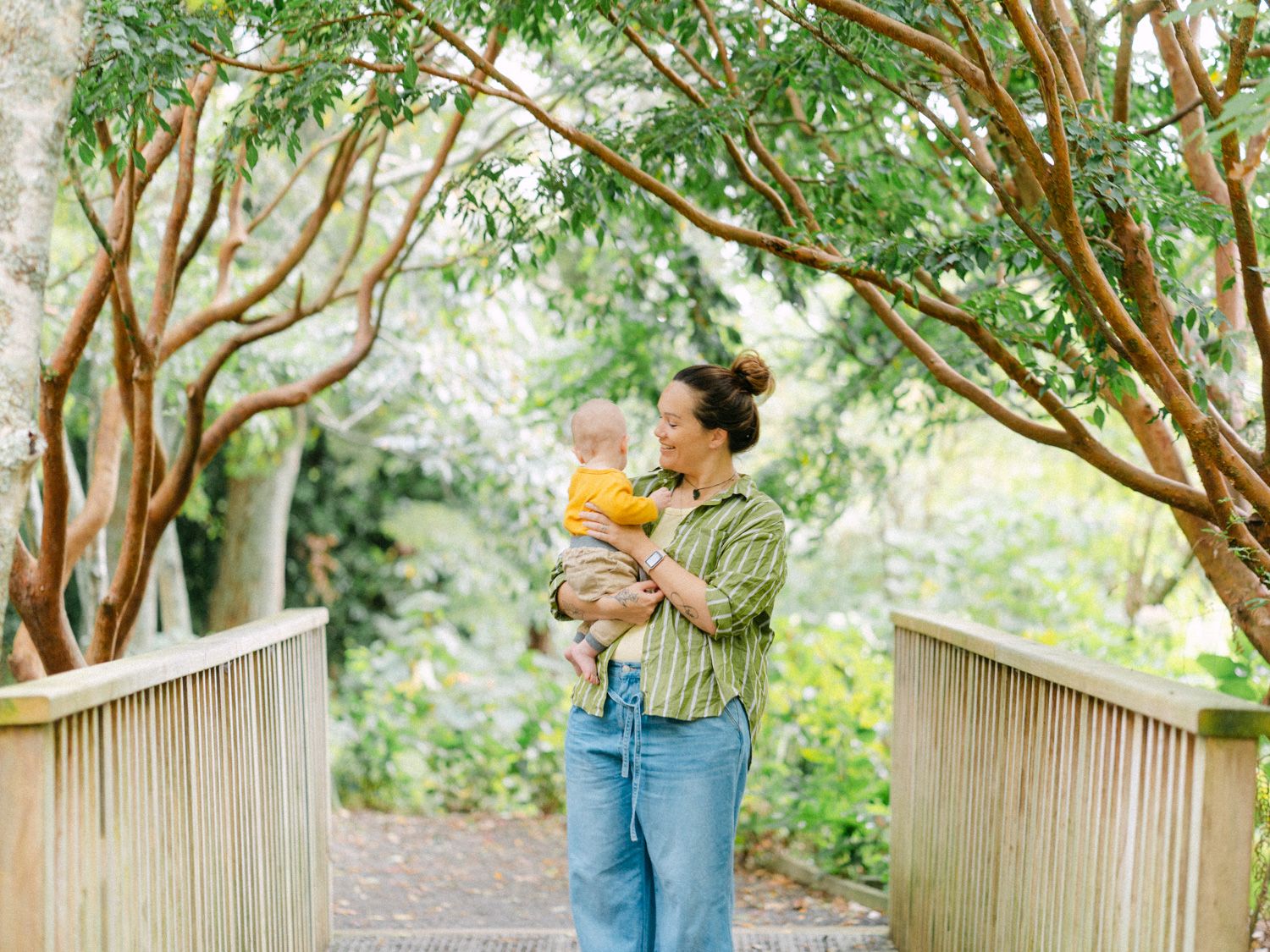 Mother in green shirt holds baby in yellow outfit on wooden bridge path surrounded by lush green trees.