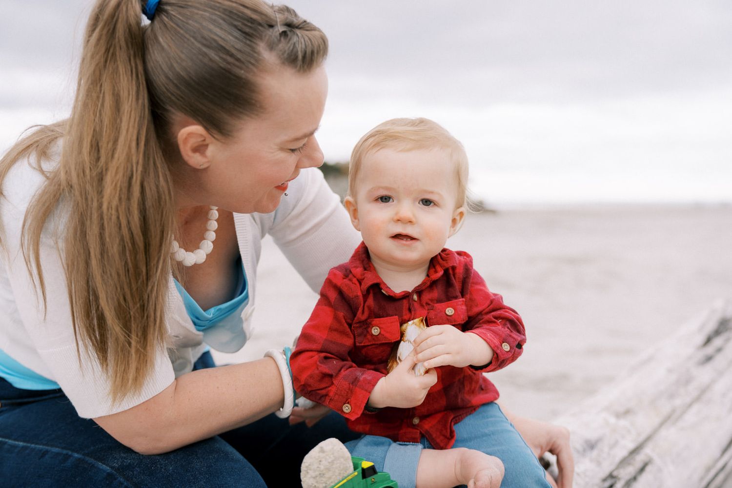 A young mother sits with her toddler in a red shirt on a sandy beach, sharing a tender moment outdoors.