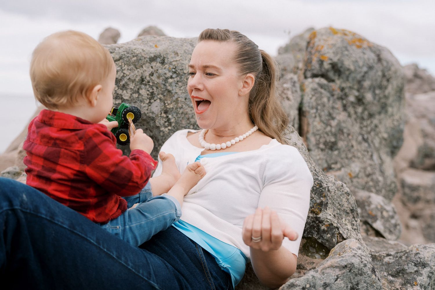 A mother laughs joyfully while her baby plays with a toy tractor outdoors among rocky terrain.