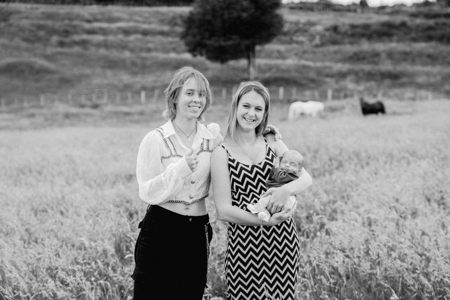 Two women smiling in a field, one holding a baby, with cattle grazing in the background, black and white photo.