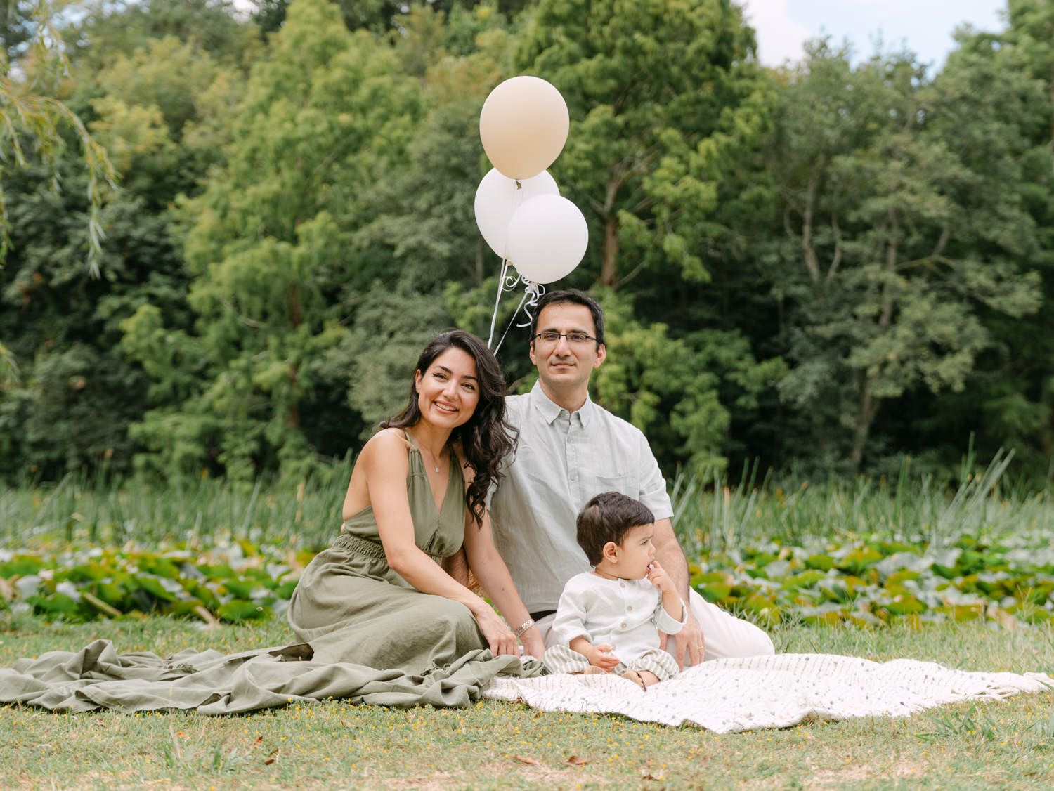 Family of three sitting on a blanket outdoors, holding balloons, smiling together in a lush green park setting.
