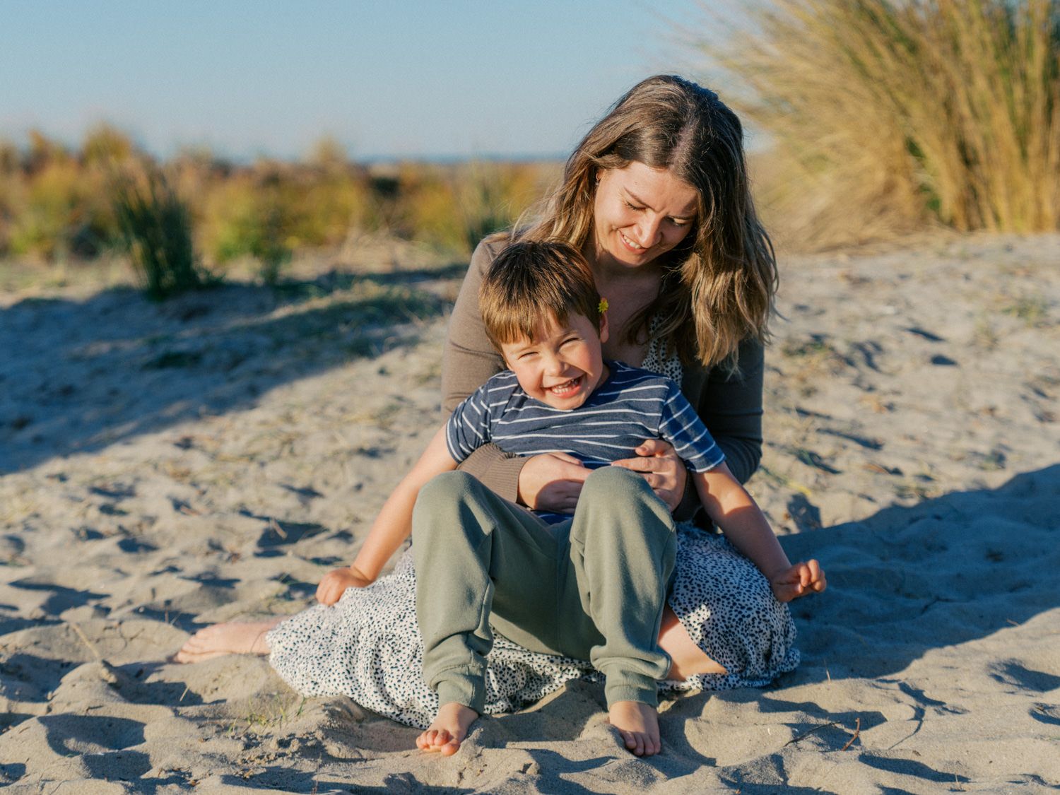 A mother and young son sit together on sandy beach dunes, smiling in warm golden sunlight.