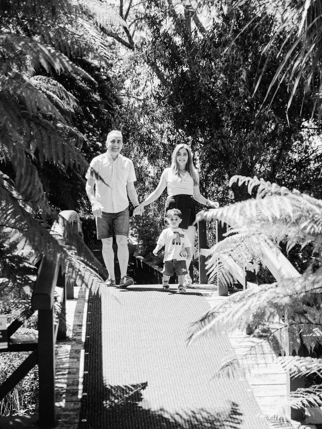 Black and white photo of a family with a toddler walking on a wooden bridge surrounded by lush fern trees.