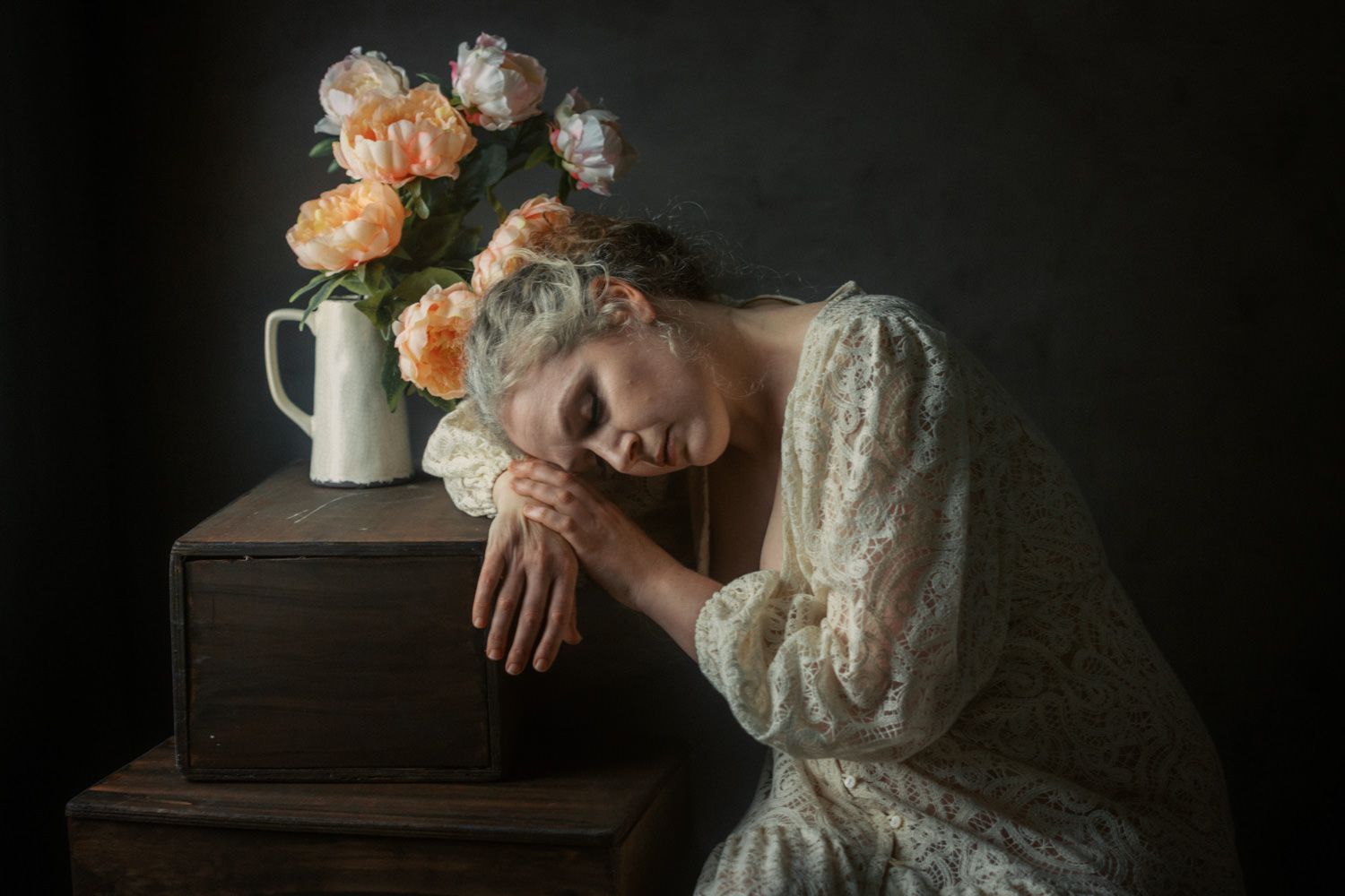 Woman in white lace dress rests head on wooden box beside peach peonies in white vase against dark background.