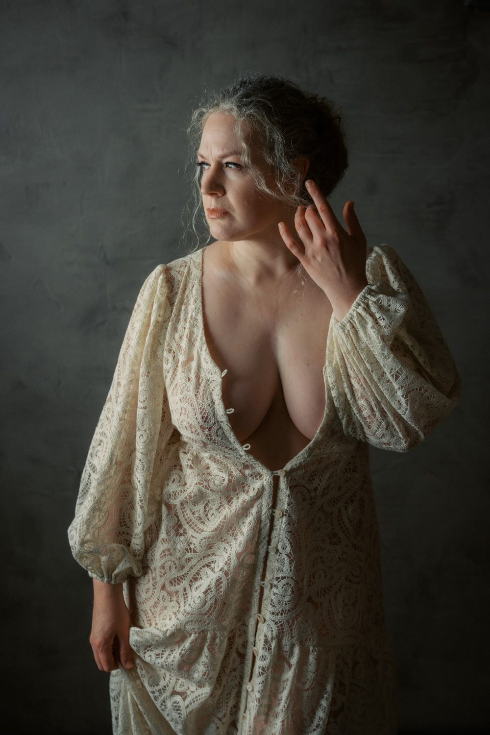 Woman in elegant cream lace dress with deep V-back, posing gracefully against dark studio background.