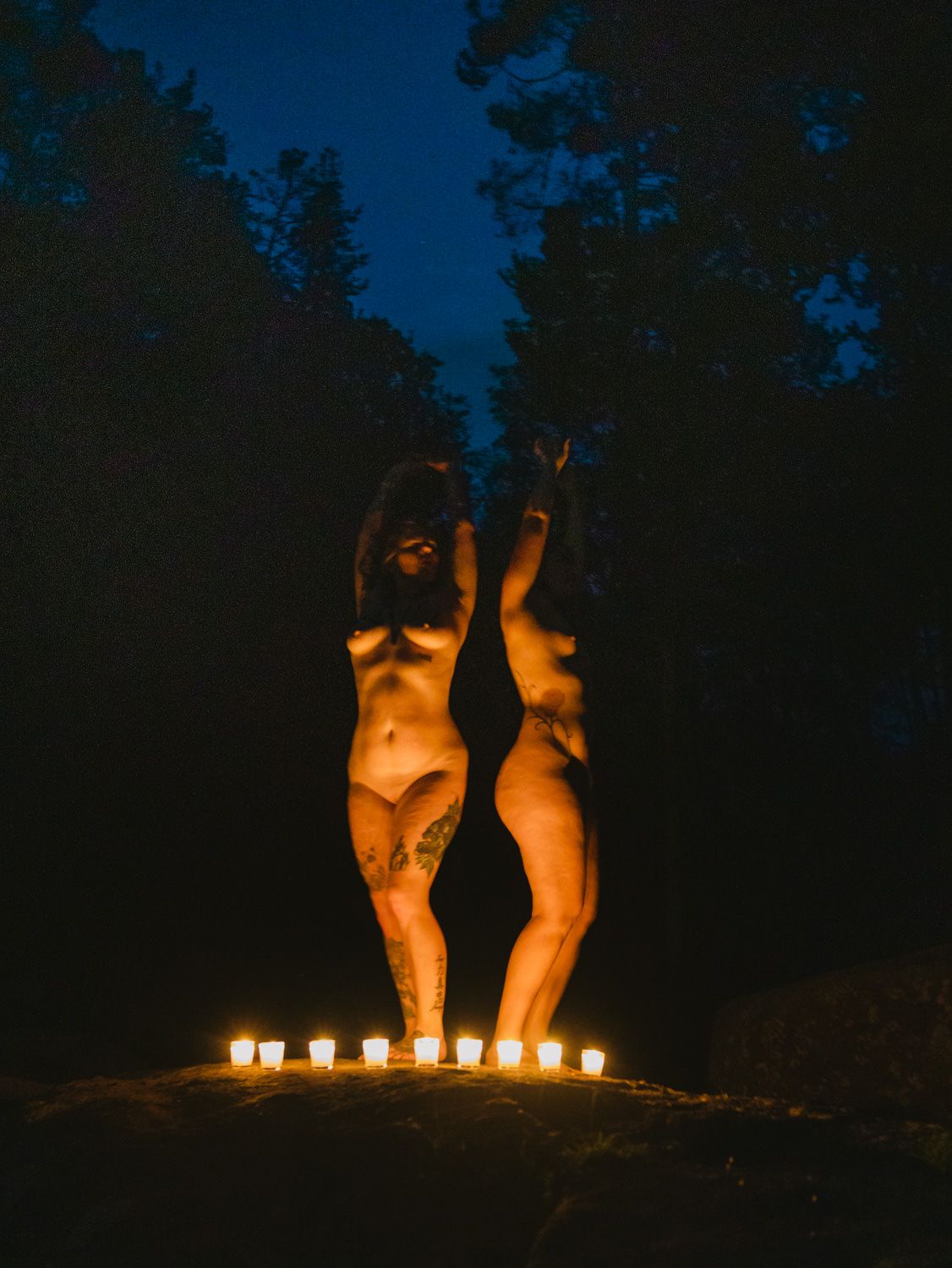 Two figures posed with arms raised surrounded by candles at night in a dark forest setting.