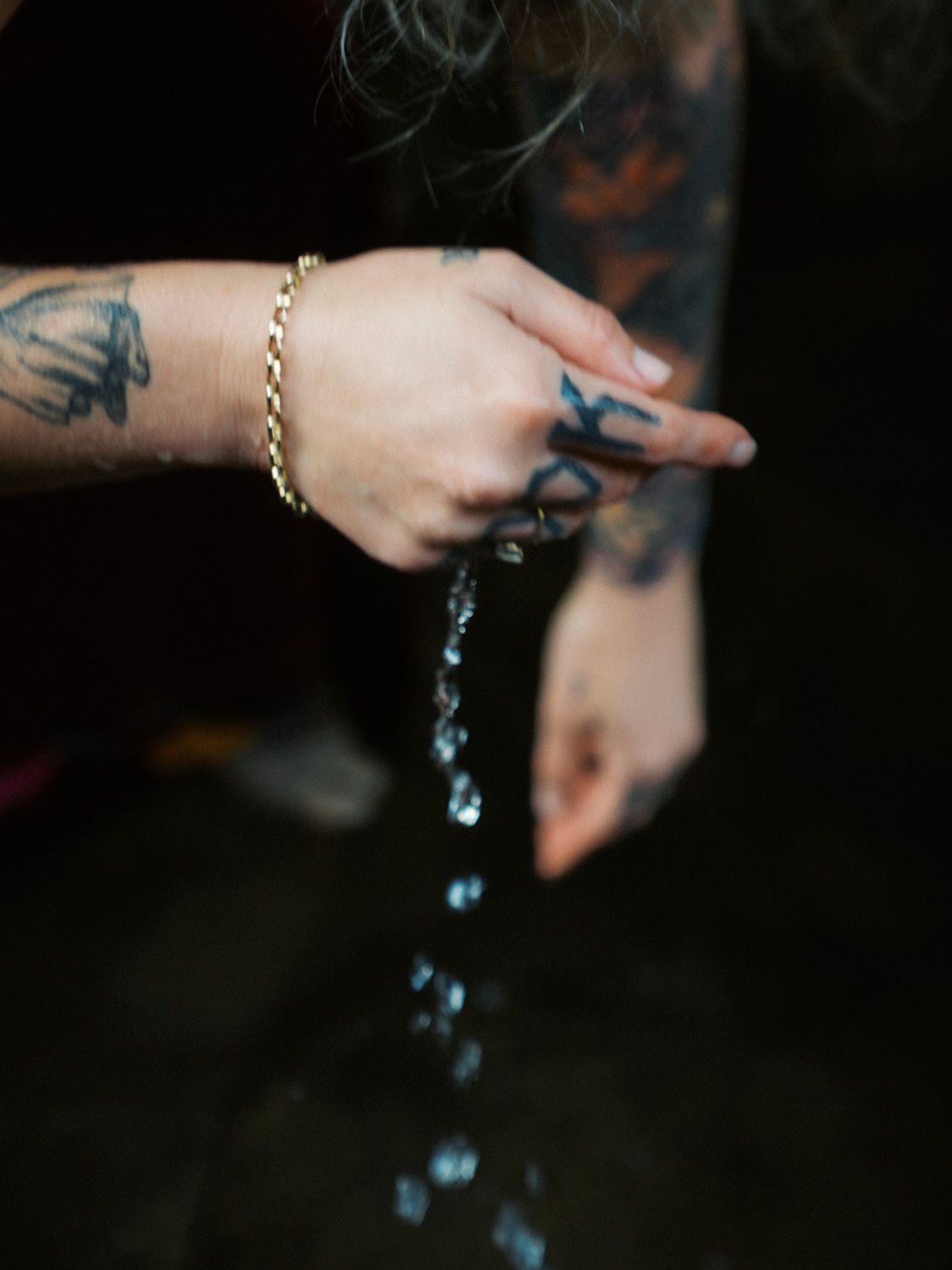 Tattooed hands with gold bracelet letting water stream through fingers into a dark reflective surface.