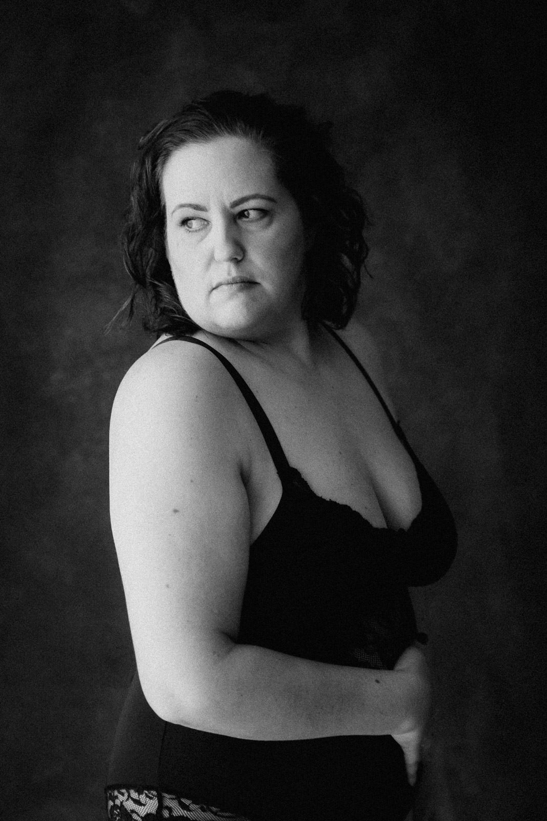 Black and white studio portrait of a confident plus-size woman in black lingerie with arms crossed.