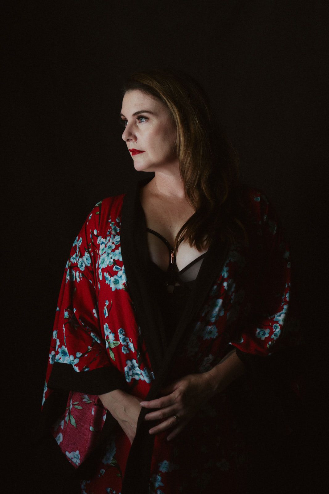 Woman in red floral kimono robe over black lingerie poses against dark background in dramatic boudoir portrait.