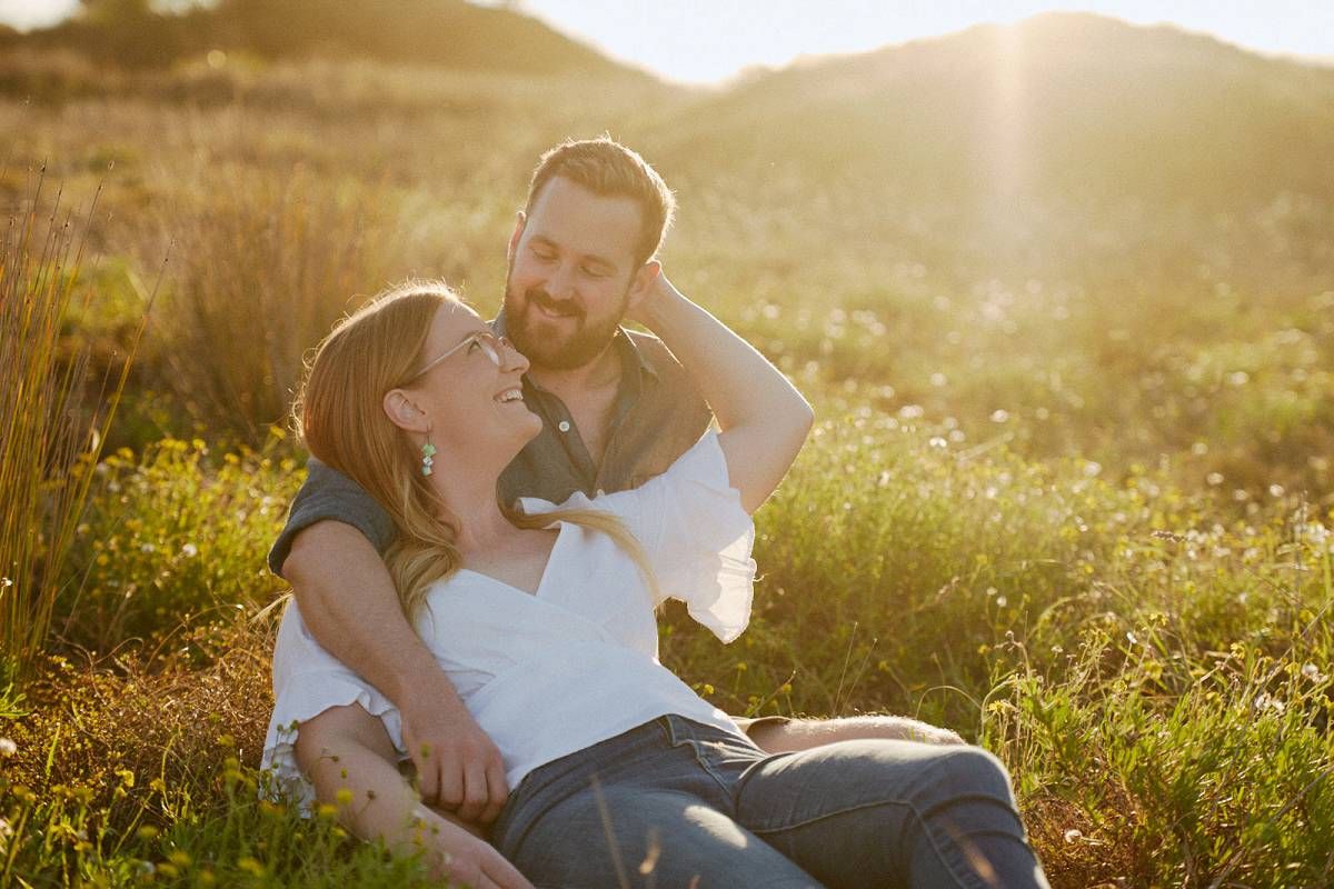 Couple relaxing in golden sunlit field, woman laughing as man gazes at her tenderly in warm sunset light.