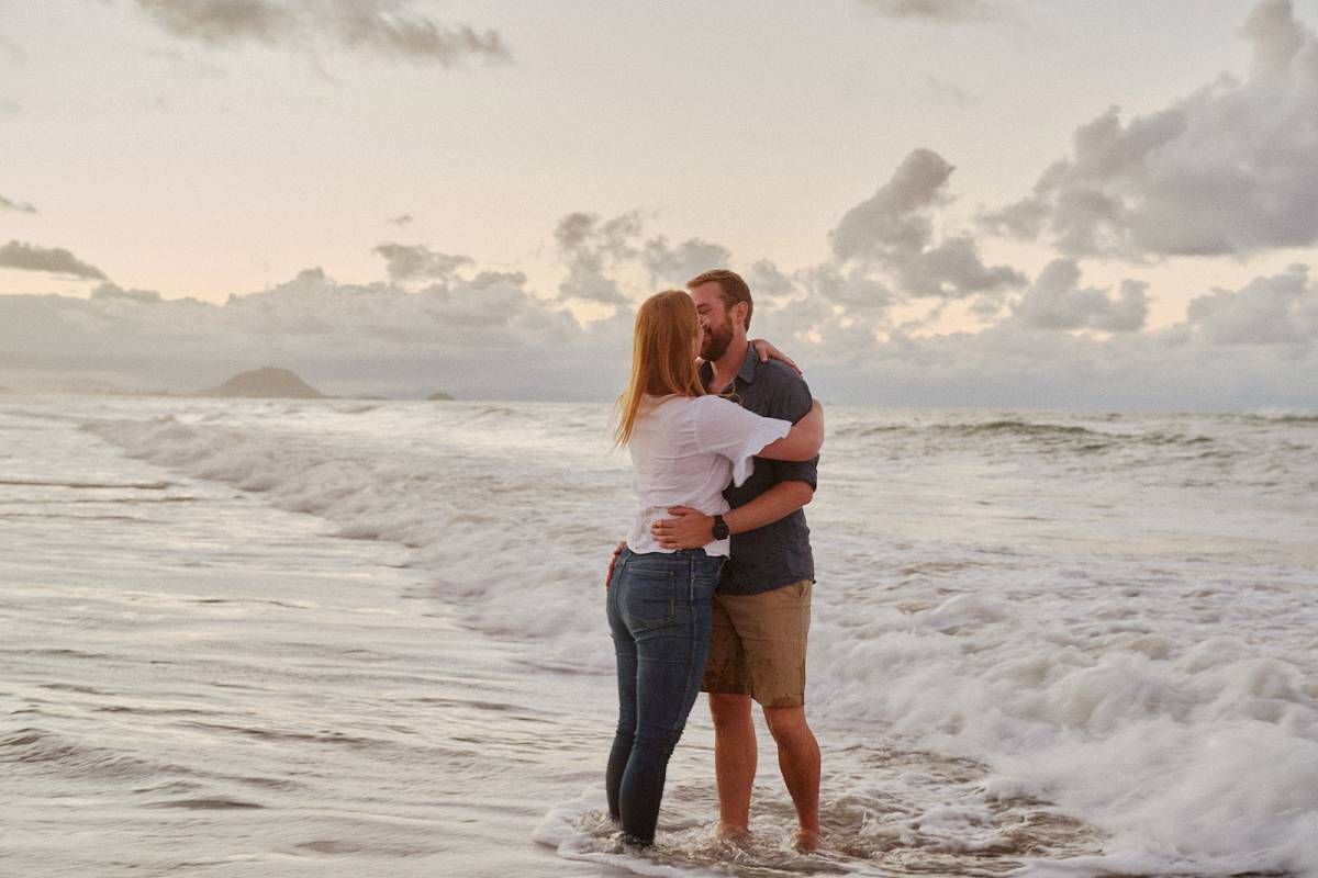 Couple kissing at the beach shore with ocean waves crashing around their feet at sunset.