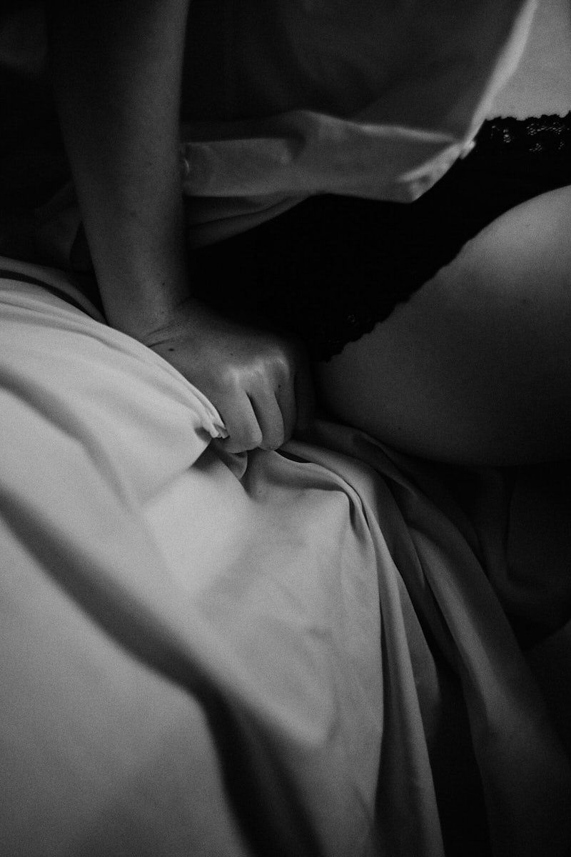 Black and white photo of a person gripping white bed sheets while lying in bed wearing black lace lingerie.