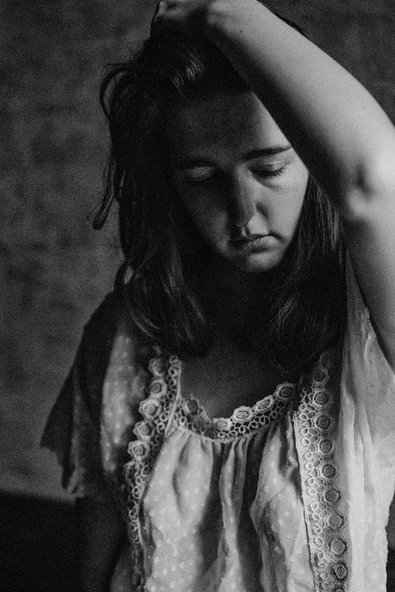 Black and white portrait of a young girl in a lace dress, arm raised, eyes closed, looking downward.