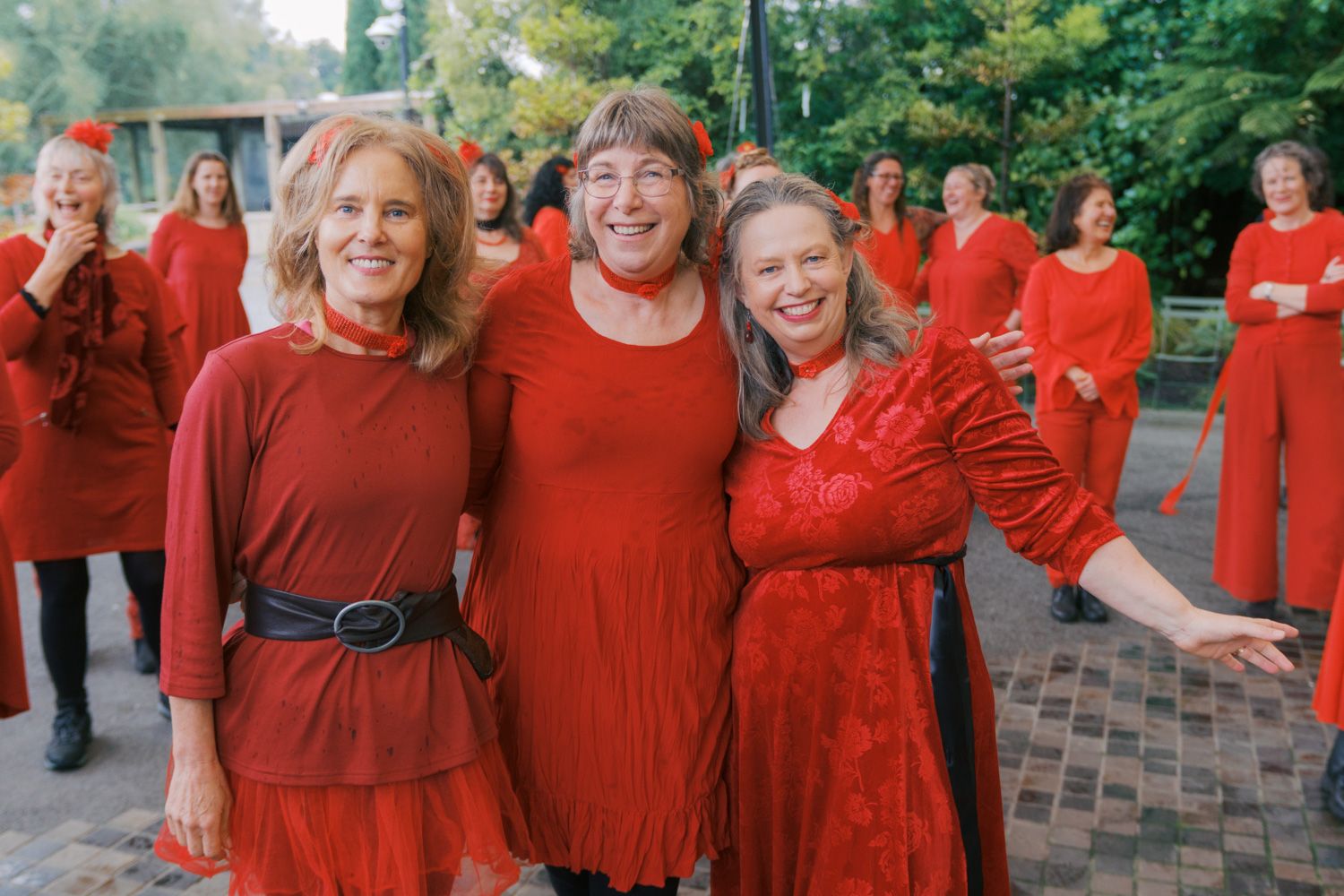 Three smiling women in red outfits pose together outdoors, with a group of people dressed in red visible in the background.