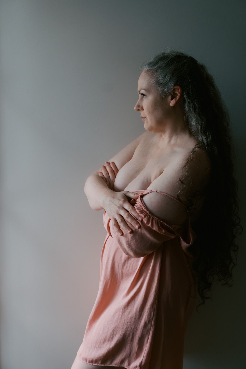 Mature woman with long gray hair in a pink dress, arms crossed, posing in profile against a gray wall.
