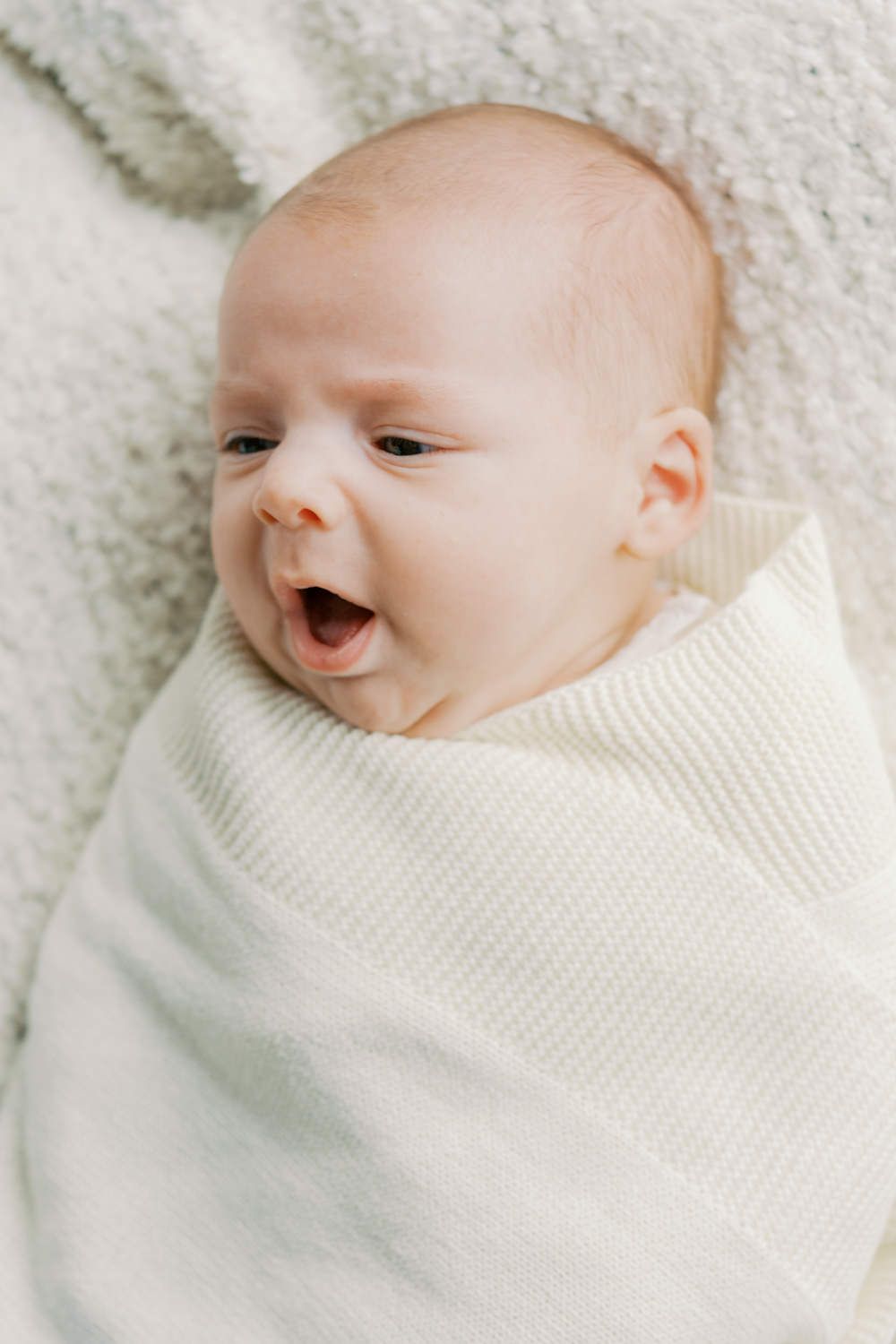 Adorable newborn baby swaddled in a white knit blanket, mouth open in a yawn, lying on a soft white surface.