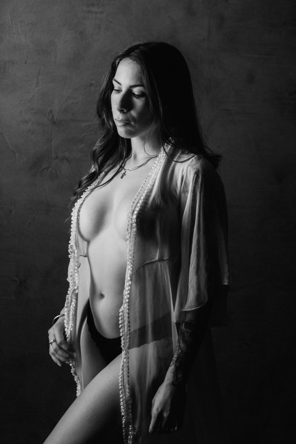 Black and white boudoir portrait of a woman in sheer robe with pearl necklace posing against dark background.
