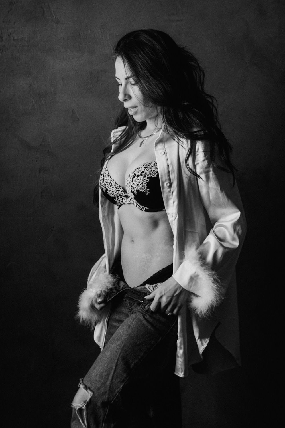 Black and white photo of a woman in lingerie, open white shirt, and ripped jeans posing in a studio.