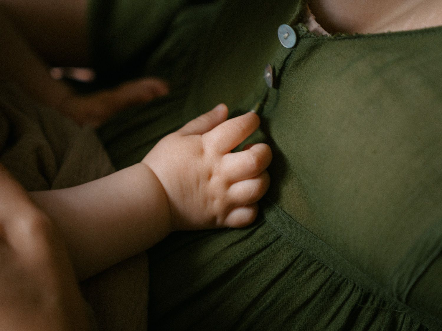 Close-up of a baby's tiny hand resting on an adult's green linen dress with buttons.