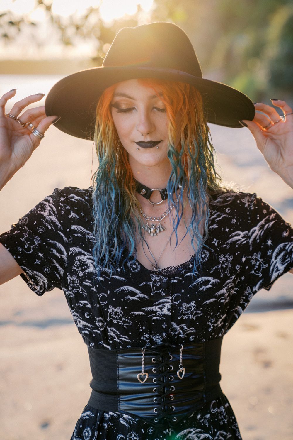 Woman with orange and blue hair wearing black wide-brim hat, dark lipstick, corset belt, and gothic patterned top outdoors.